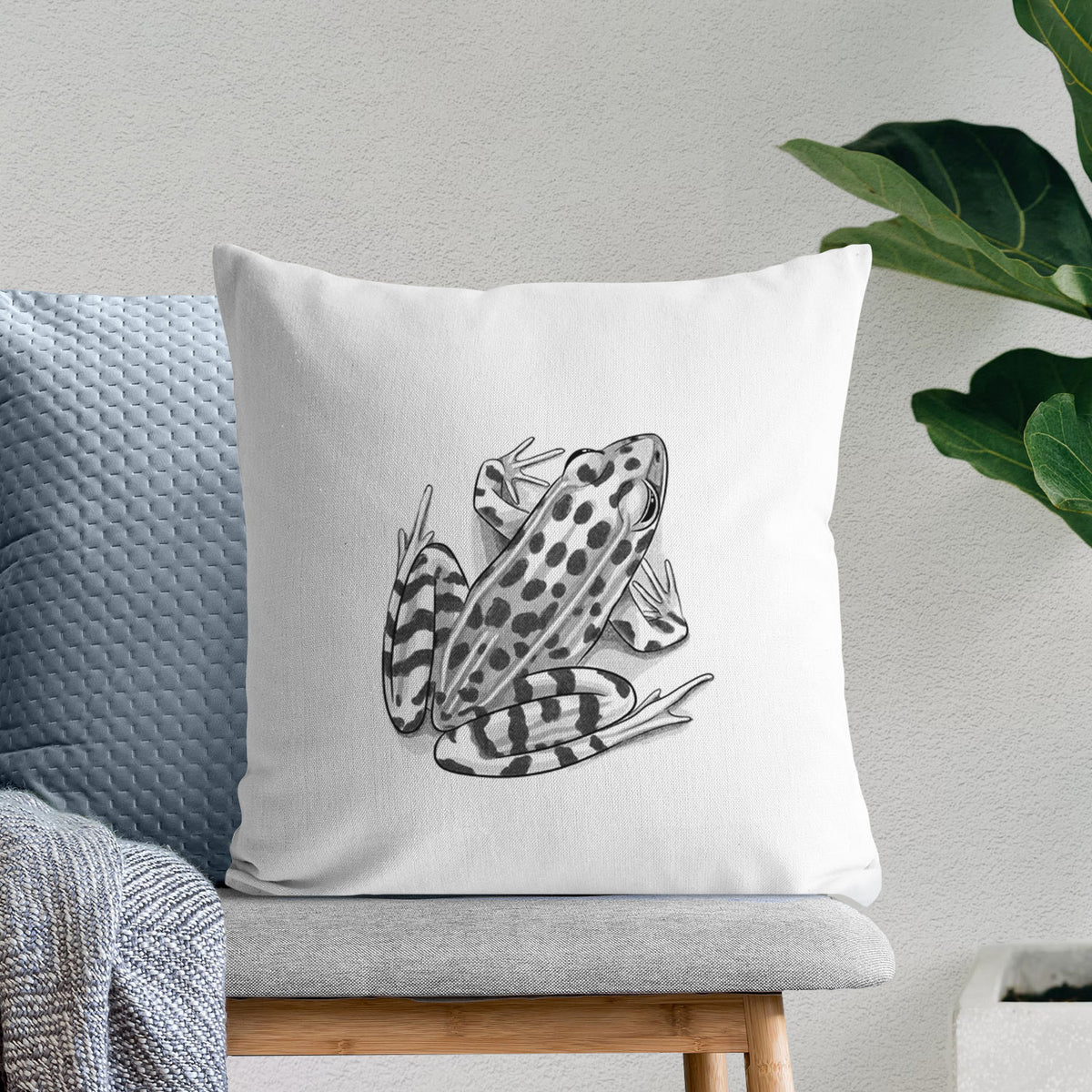 White pillow cover printed with a detailed black ink illustration of a spotted northern leopard frog, on a bench beside plants.