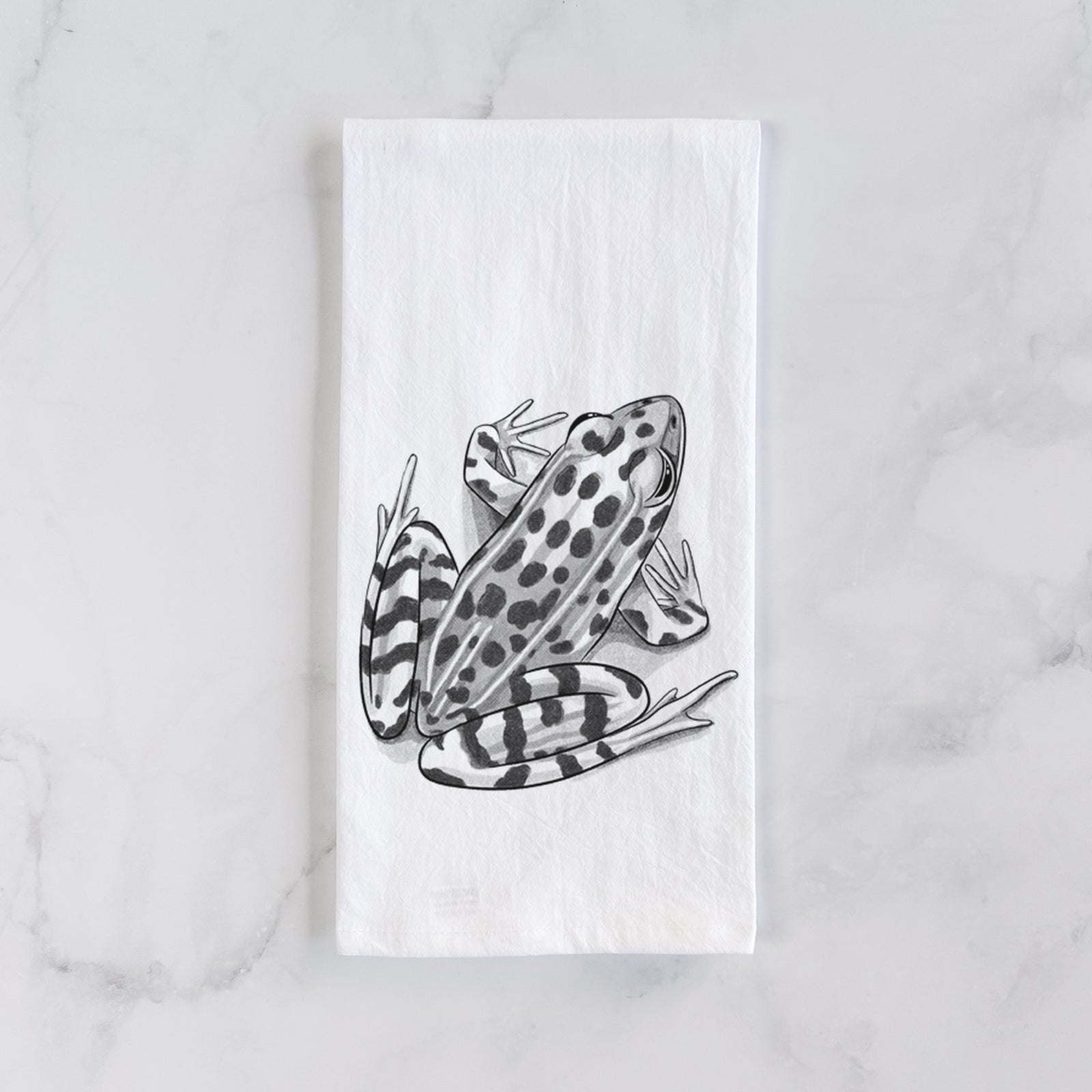 White cotton tea towel printed with a detailed black ink illustration of a spotted northern leopard frog, on a marble counter beside a round wooden cutting board and greenery.