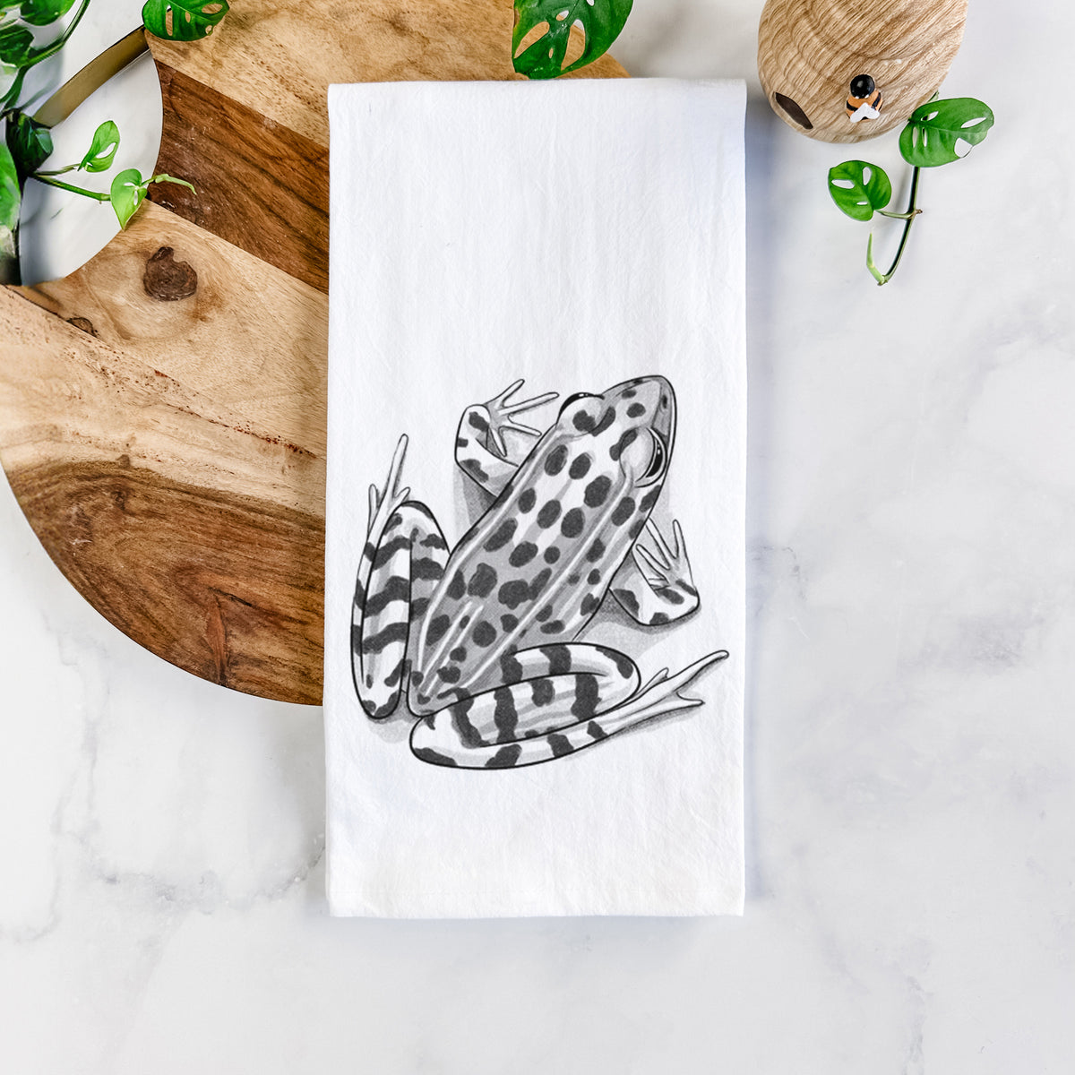 White cotton tea towel printed with a detailed black ink illustration of a spotted northern leopard frog, on a marble counter beside a round wooden cutting board and greenery.