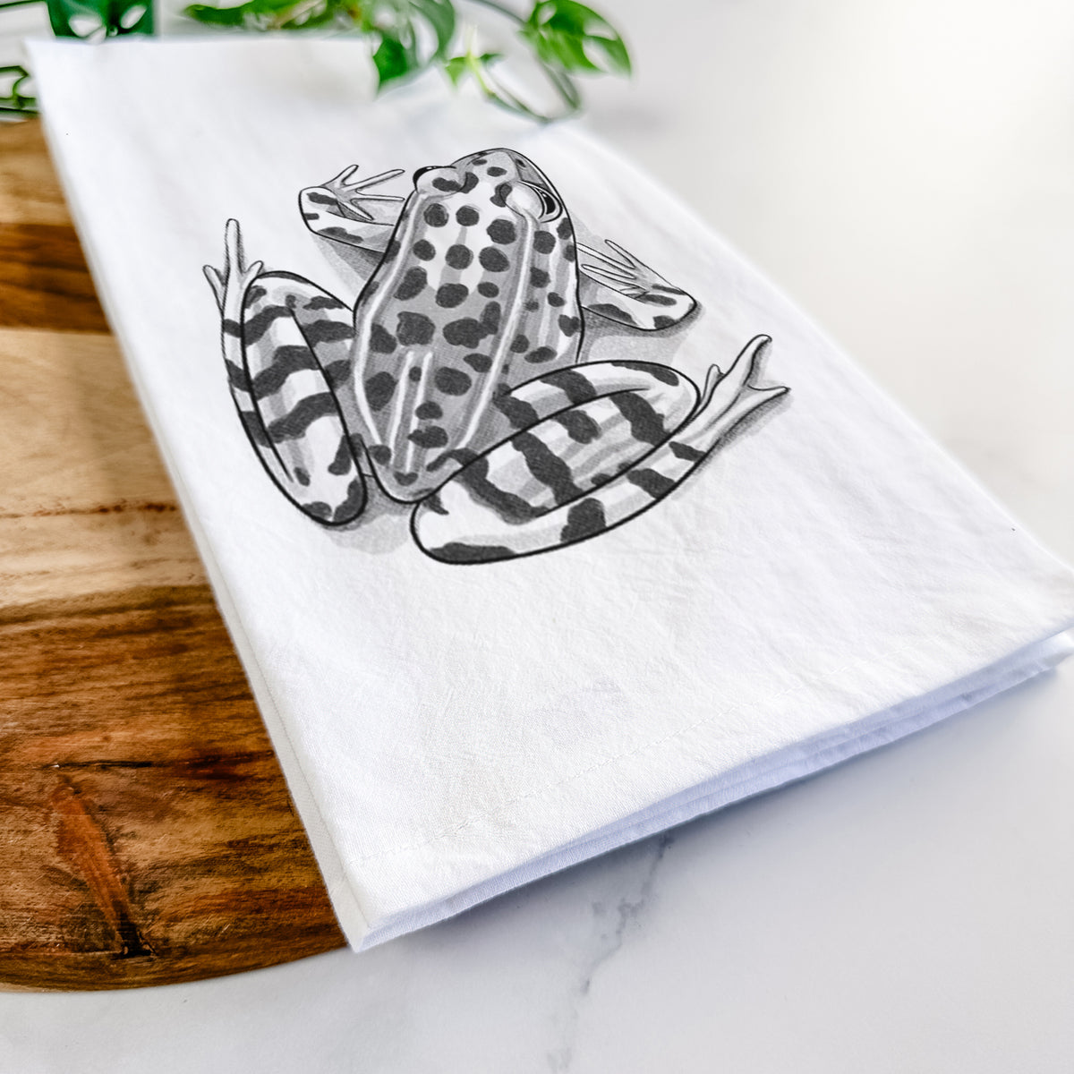 Close-up of a folded white tea towel on a wooden board, showing a detailed black ink illustration of a spotted northern leopard frog.