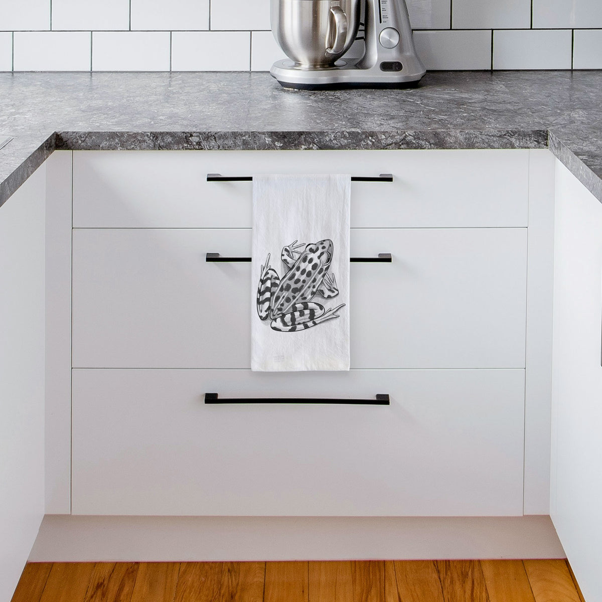 White tea towel printed with a detailed black ink illustration of a spotted northern leopard frog, hanging from a drawer handle in a bright white kitchen.