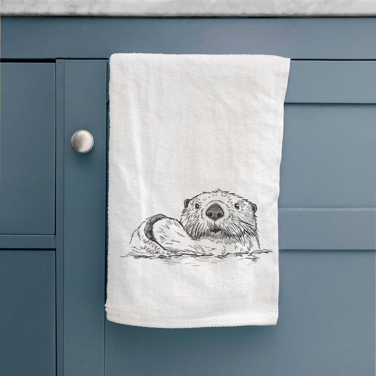 White hand towel printed with a black ink illustration of a northern river otter peeking out of the water, hanging on a cabinet door.