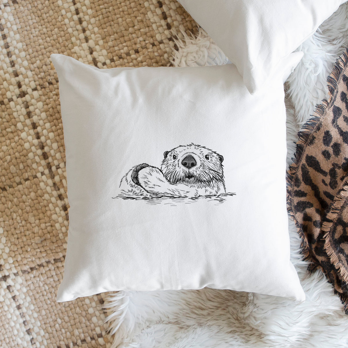 White pillow cover printed with a black ink illustration of a northern river otter peeking out of the water, on a woven chair.