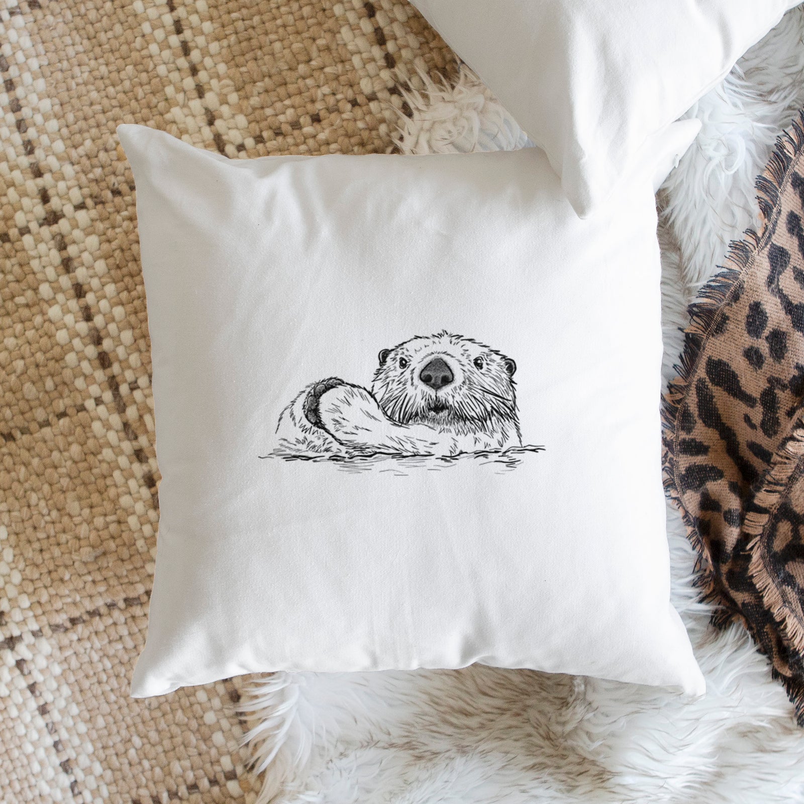 White pillow cover printed with a black ink illustration of a northern river otter peeking out of the water, on a bench beside plants.