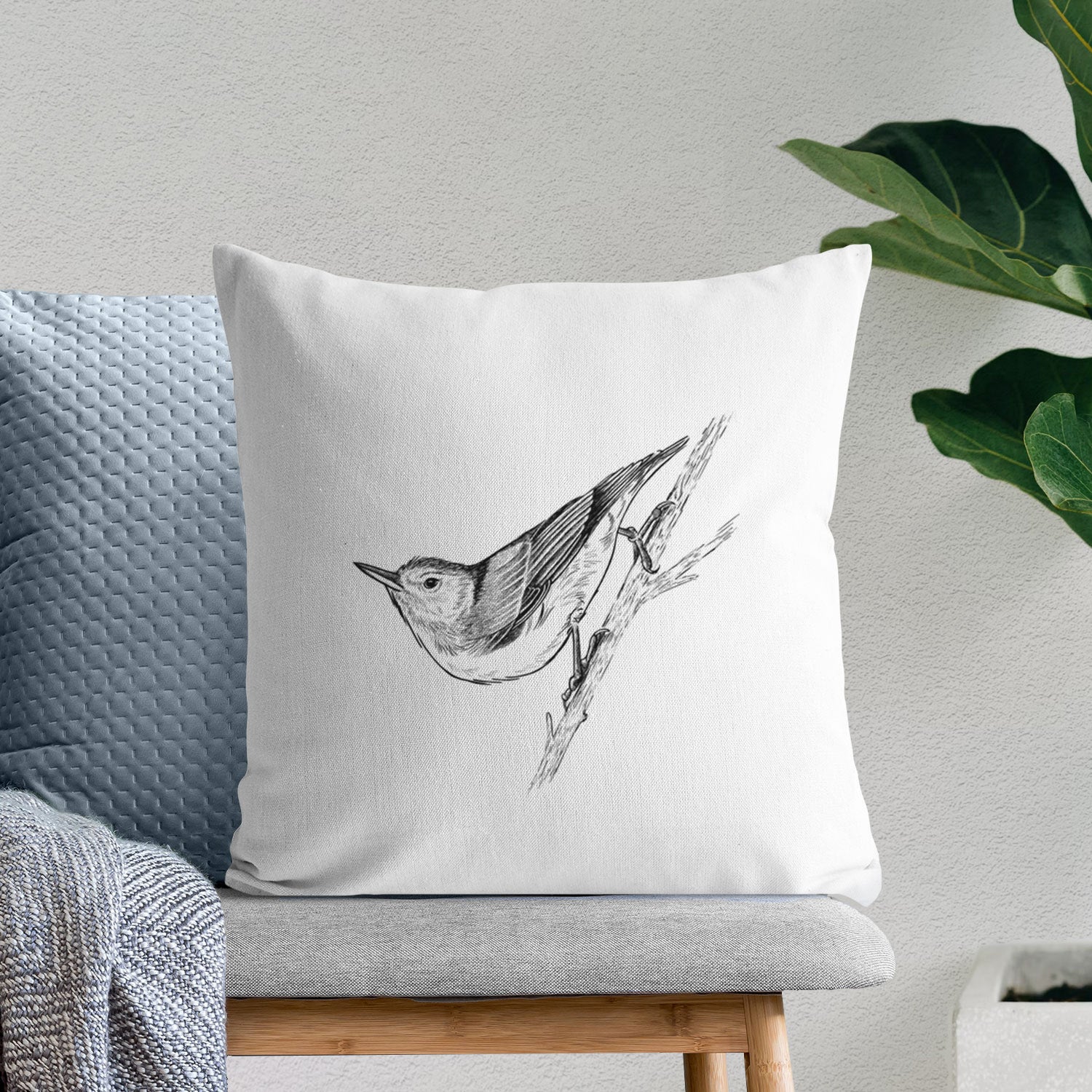 White pillow cover printed with a detailed black ink sketch of a nuthatch clinging to a branch, on a bench beside plants.
