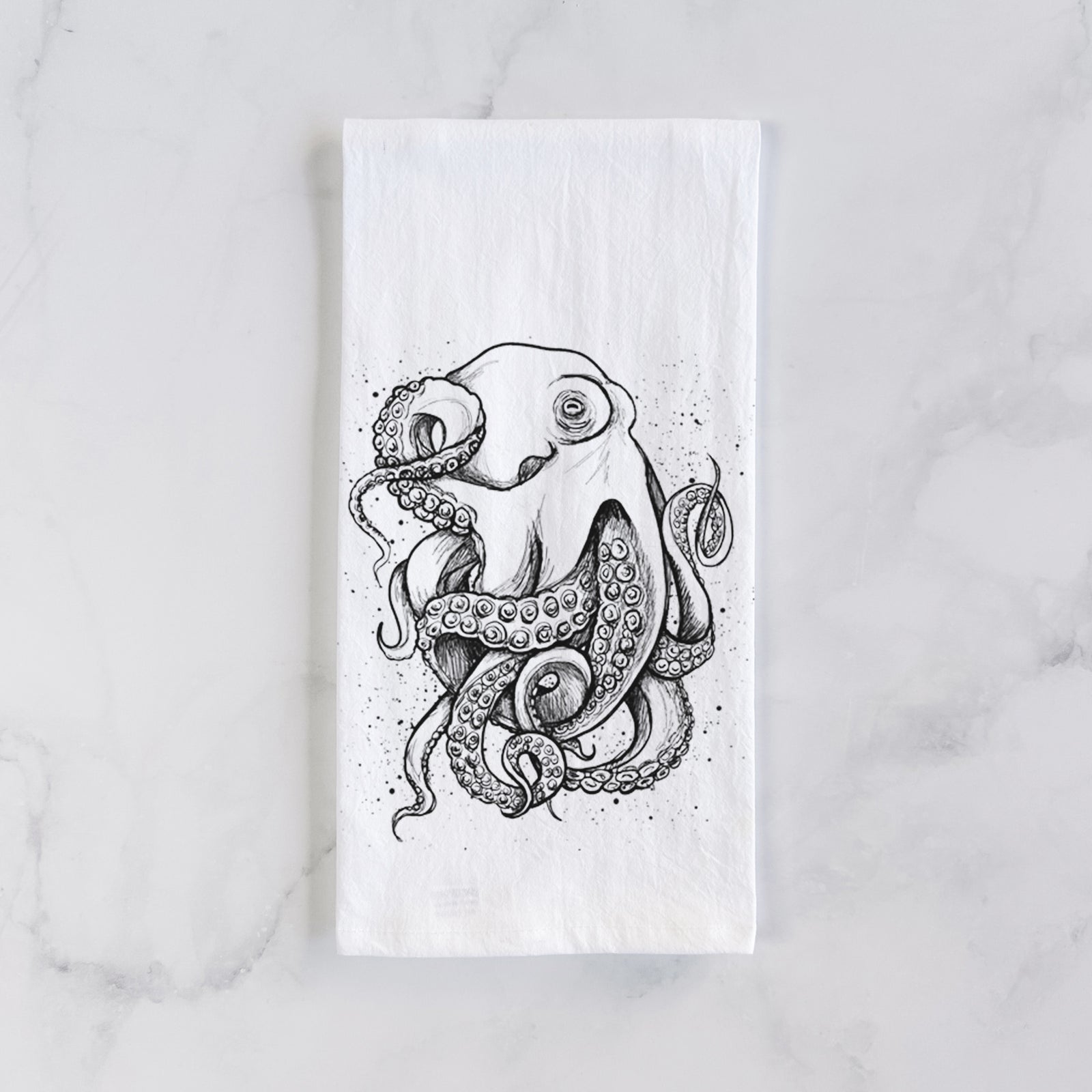 White cotton tea towel printed with a detailed black ink illustration of a common octopus with curling tentacles, on a marble counter beside a round wooden cutting board and greenery.