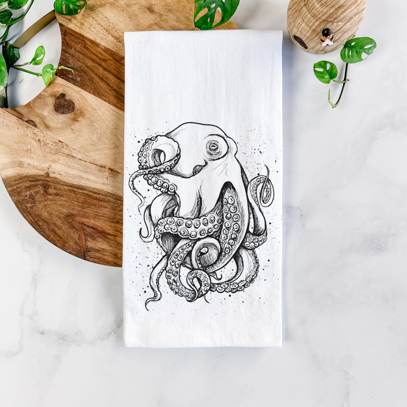 White cotton tea towel printed with a detailed black ink illustration of a common octopus with curling tentacles, on a marble counter beside a round wooden cutting board and greenery.