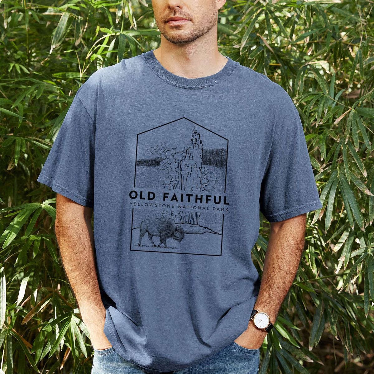 Man wearing a Comfort Colors t-shirt printed with a black line art badge of the Old Faithful geyser erupting with a bison nearby, with the words Old Faithful, Yellowstone National Park, outdoors.
