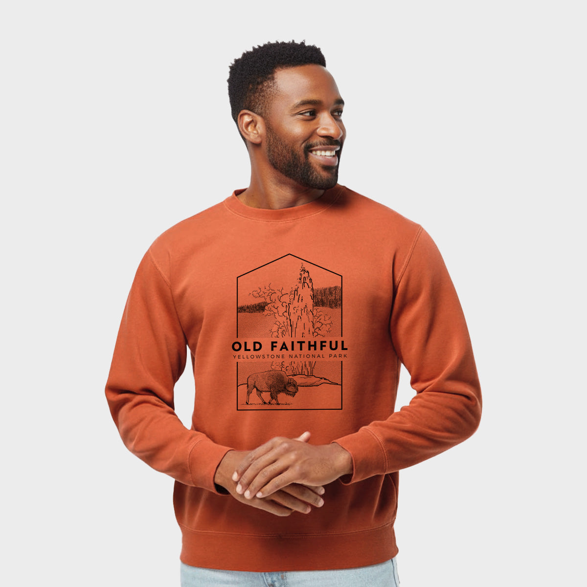 Model wearing an amber orange pigment dyed crewneck sweatshirt printed with a black line art badge of the Old Faithful geyser erupting with a bison nearby, with the words Old Faithful, Yellowstone National Park.