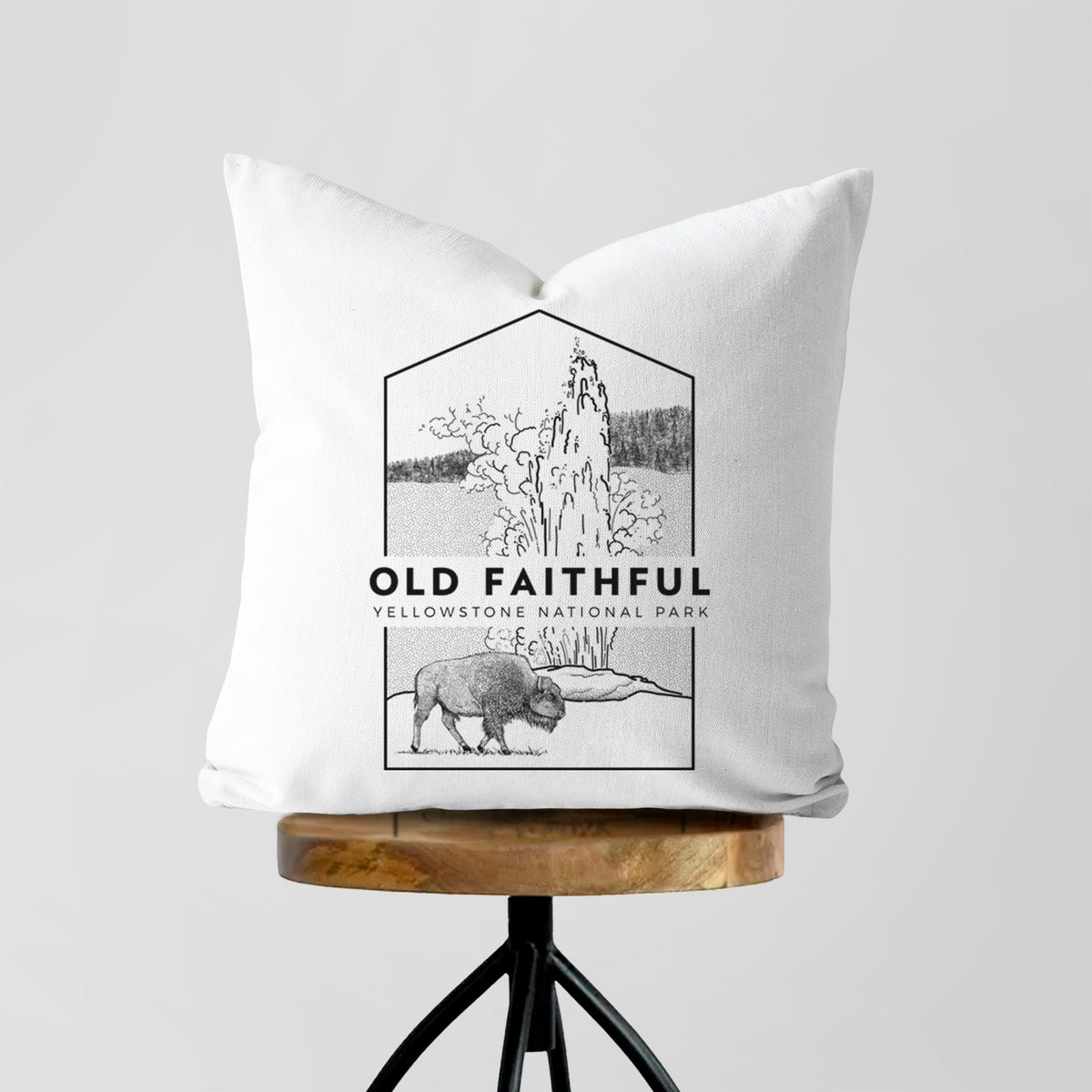 White pillow cover printed with a black line art badge of the Old Faithful geyser erupting with a bison nearby, with the words Old Faithful, Yellowstone National Park, on a wooden stool.