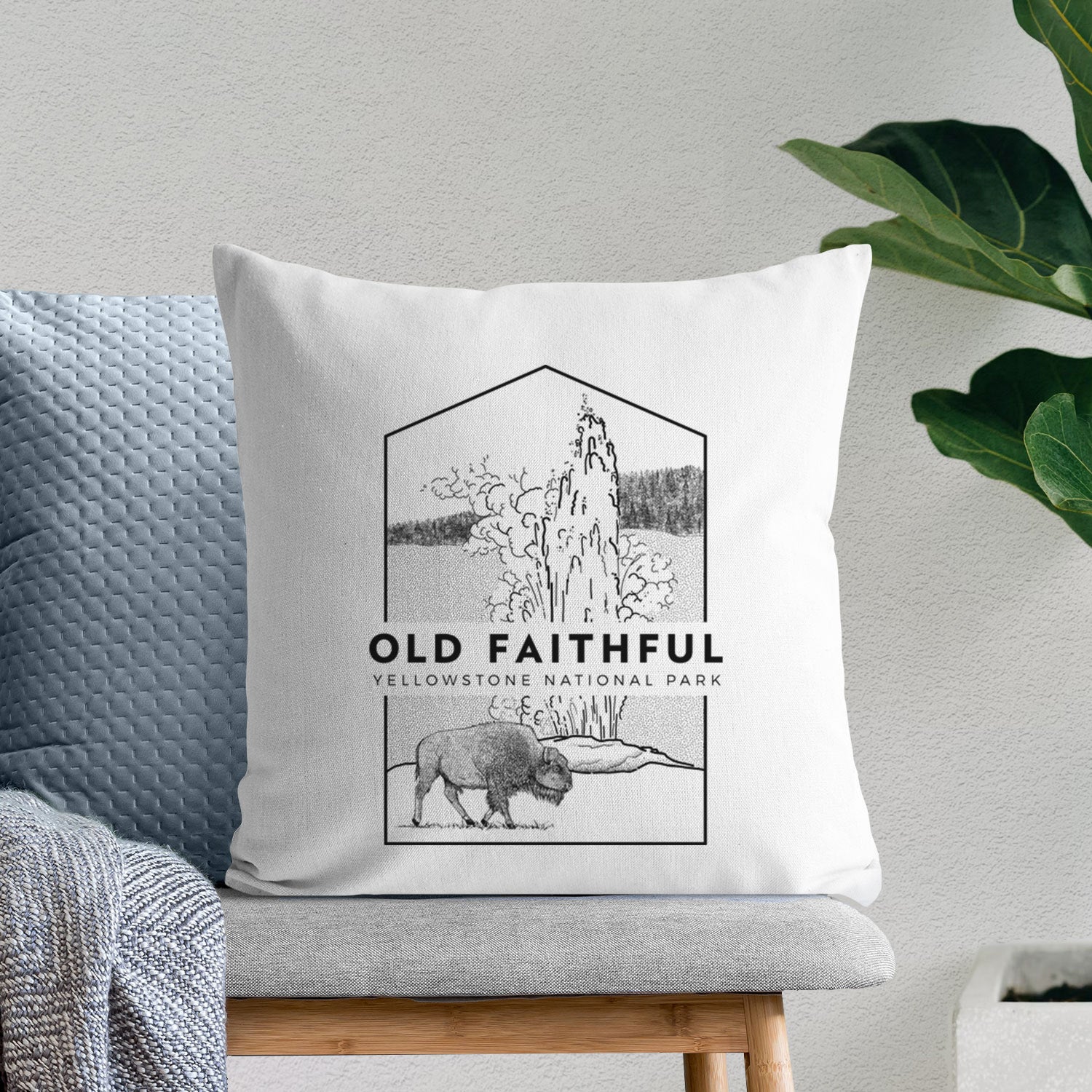 White pillow cover printed with a black line art badge of the Old Faithful geyser erupting with a bison nearby, with the words Old Faithful, Yellowstone National Park, on a bench beside plants.