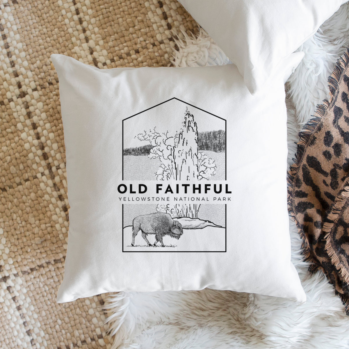 White pillow cover printed with a black line art badge of the Old Faithful geyser erupting with a bison nearby, with the words Old Faithful, Yellowstone National Park, on a woven chair.