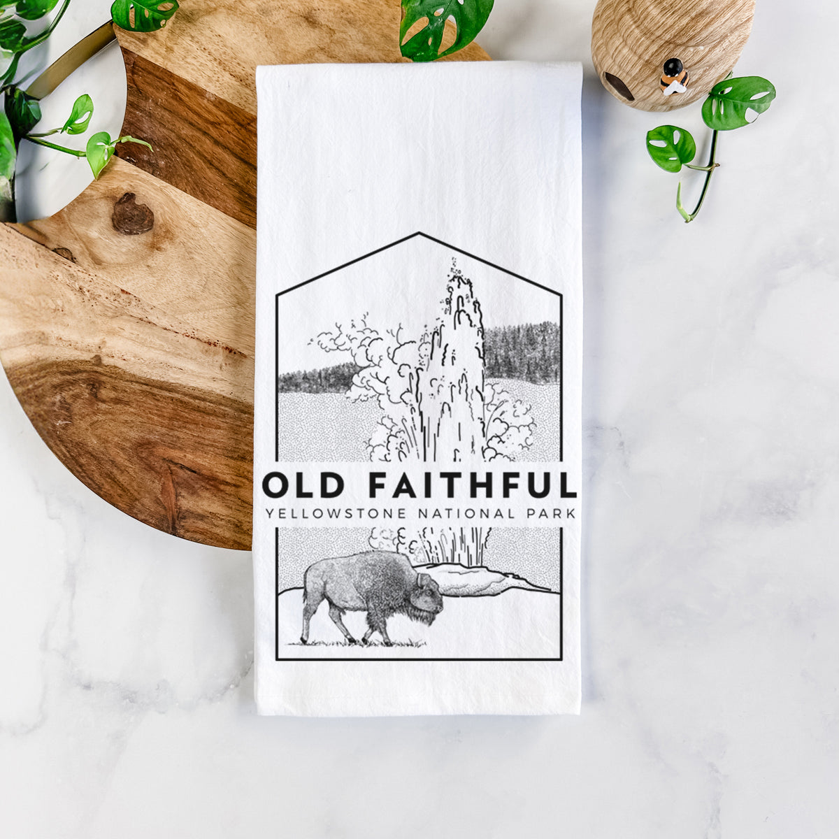 White cotton tea towel printed with a black line art badge of the Old Faithful geyser erupting with a bison nearby, with the words Old Faithful, Yellowstone National Park, on a marble counter beside a round wooden cutting board and greenery.