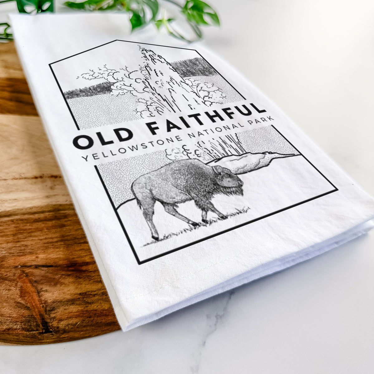 Close-up of a folded white tea towel on a wooden board, showing a black line art badge of the Old Faithful geyser erupting with a bison nearby, with the words Old Faithful, Yellowstone National Park.