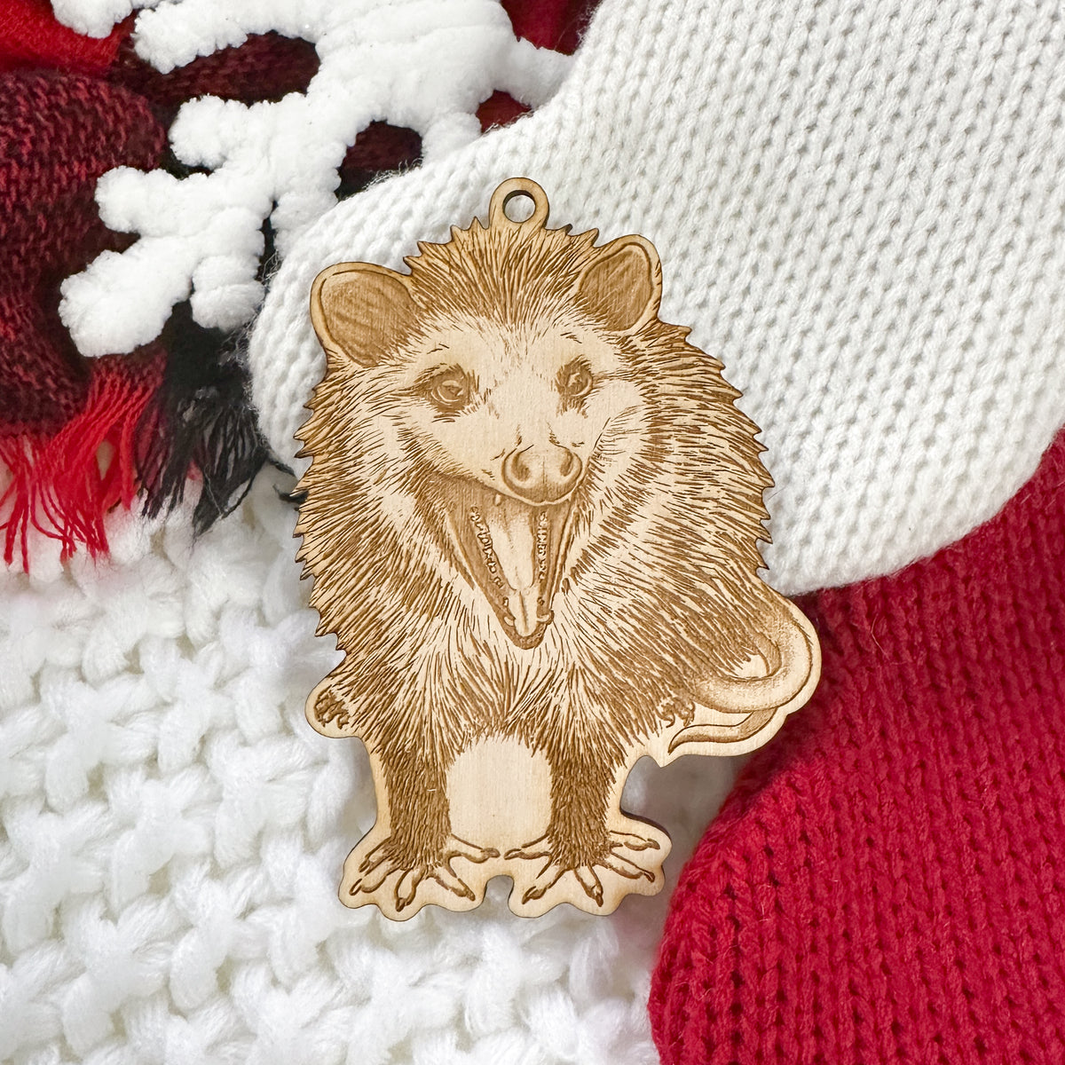 Wooden ornament featuring a detailed engraving of a hissing opossum standing and facing forward, on a knit blanket with pine cones.