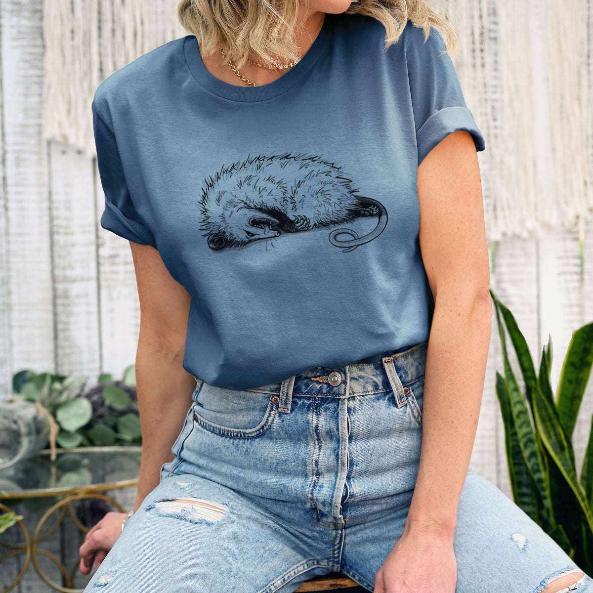 Woman wearing a steel blue lightweight cotton t-shirt printed with a black ink illustration of an opossum lying on its side playing dead.