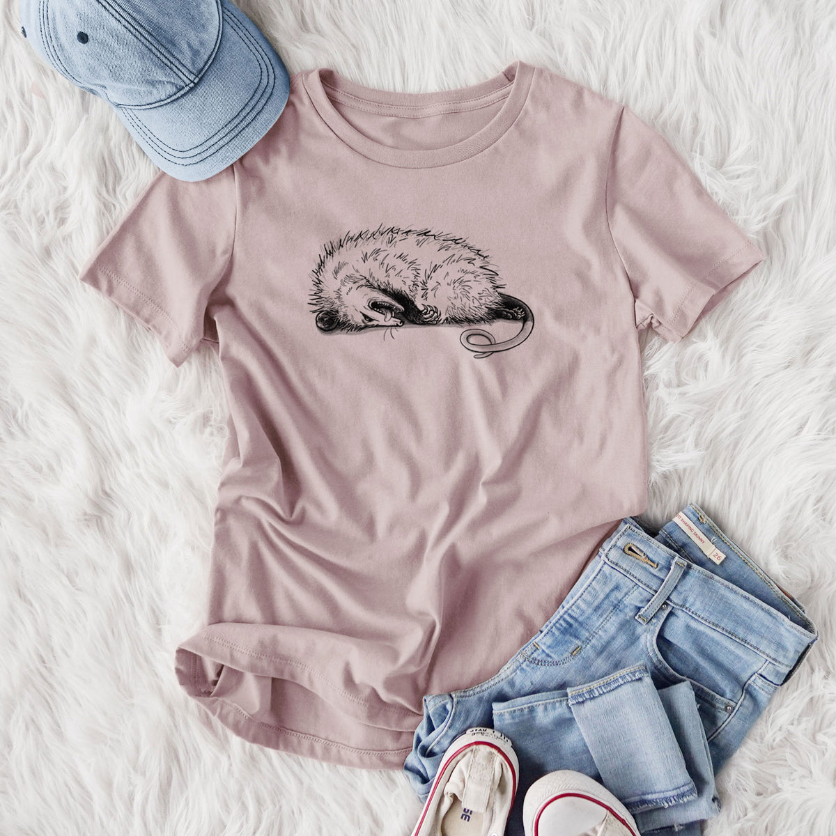 Women's relaxed fit cotton t-shirt printed with a black ink illustration of an opossum lying on its side playing dead, styled flat on a white faux fur rug with jeans.