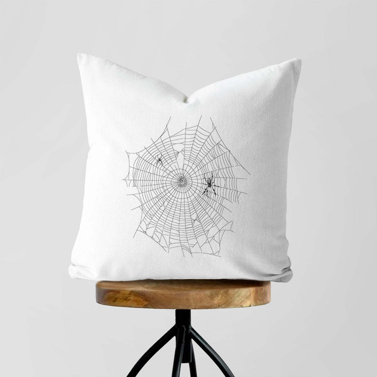 White pillow cover printed with a black ink line drawing of an orb weaver spider in the center of its web, on a wooden stool.