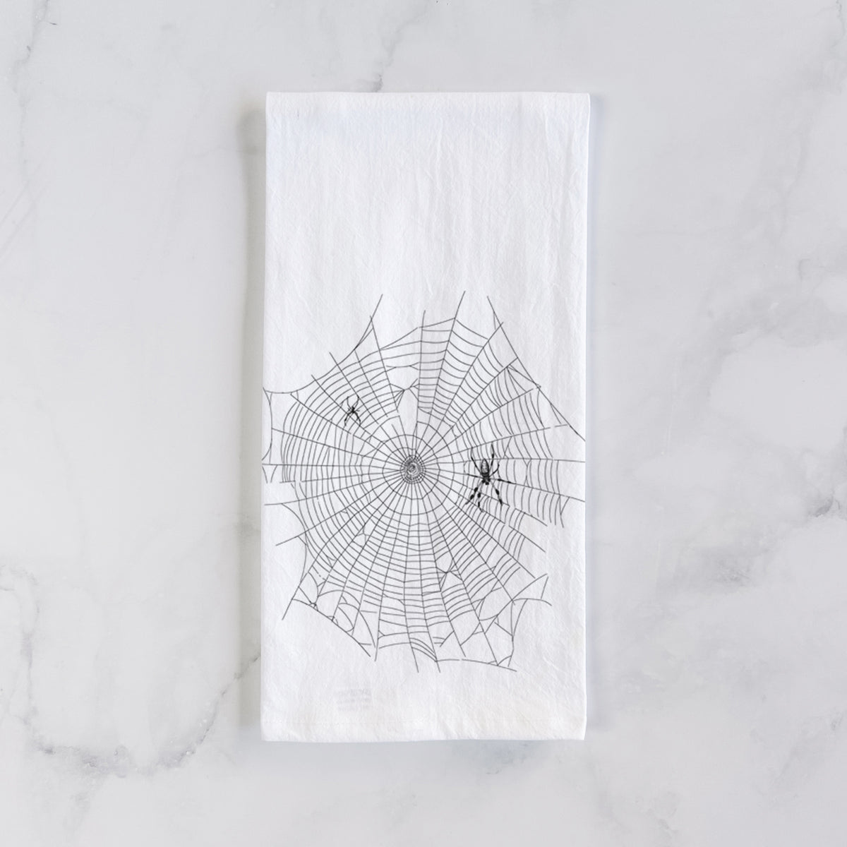 White cotton tea towel laid flat on marble, printed with a black ink line drawing of an orb weaver spider in the center of its web.