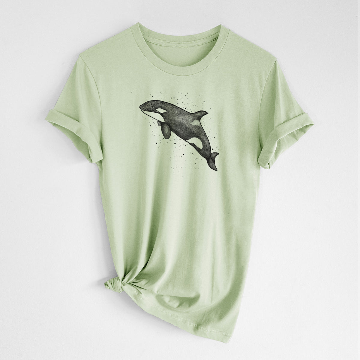 Spring green lightweight cotton t-shirt laid flat with the sleeves rolled, printed with a black ink illustration of an orca leaping, with ink splatters.