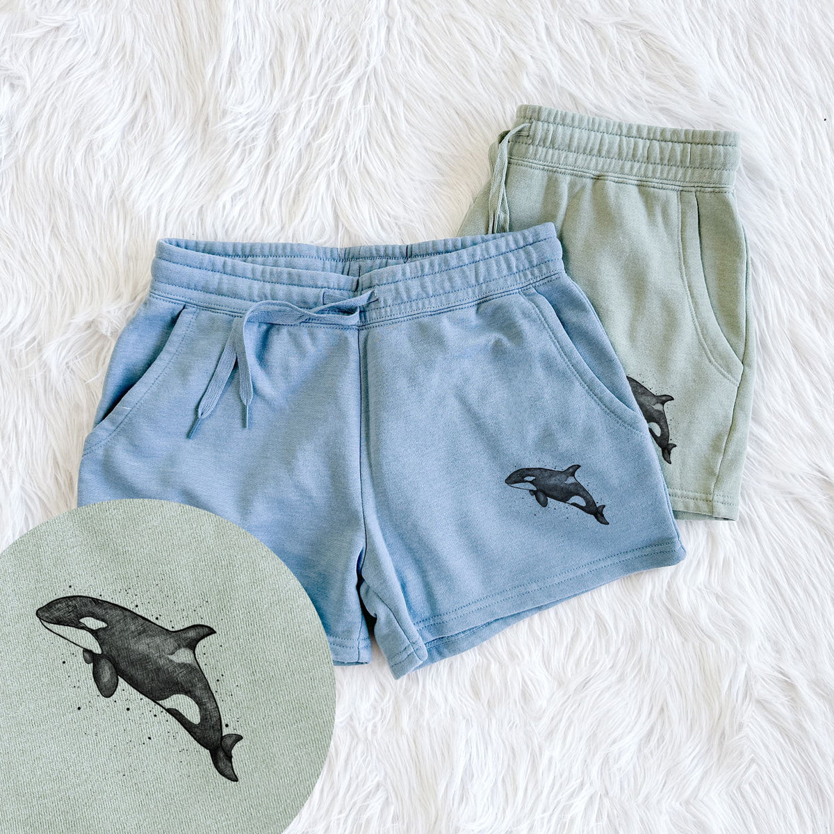 Two pairs of Cali Wave shorts in different colors printed with a black ink illustration of an orca leaping, with ink splatters, with a close-up of the print.