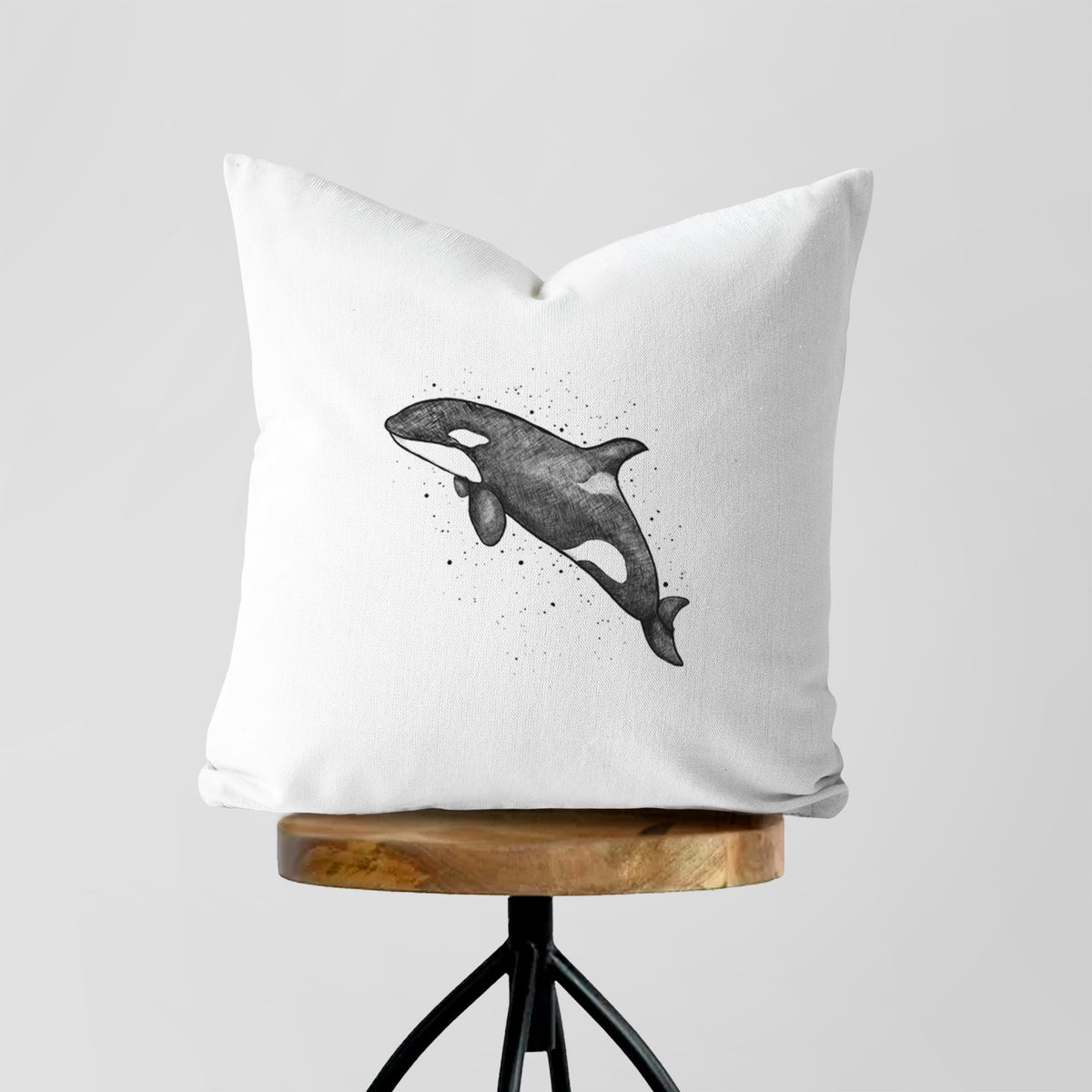 White pillow cover printed with a black ink illustration of an orca leaping, with ink splatters, on a wooden stool.