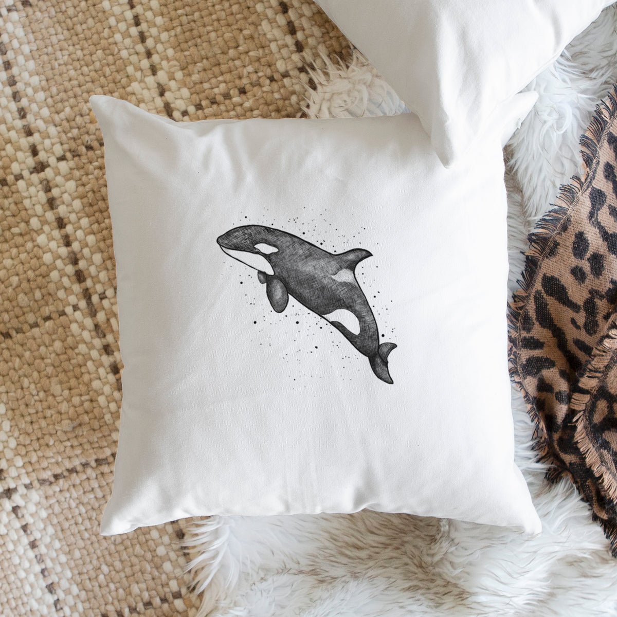 White pillow cover printed with a black ink illustration of an orca leaping, with ink splatters, on a woven chair.