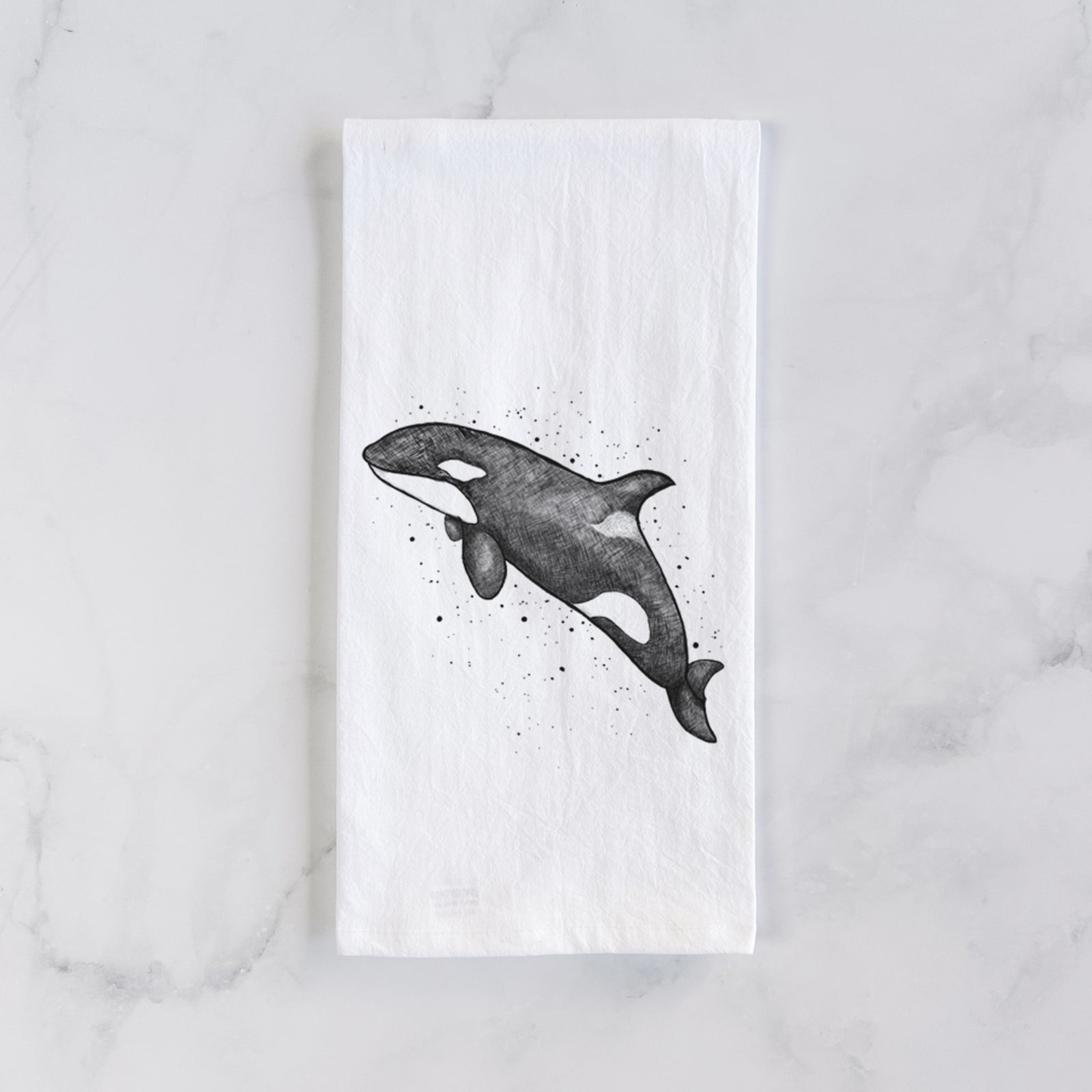 White cotton tea towel printed with a black ink illustration of an orca leaping, with ink splatters, on a marble counter beside a round wooden cutting board and greenery.
