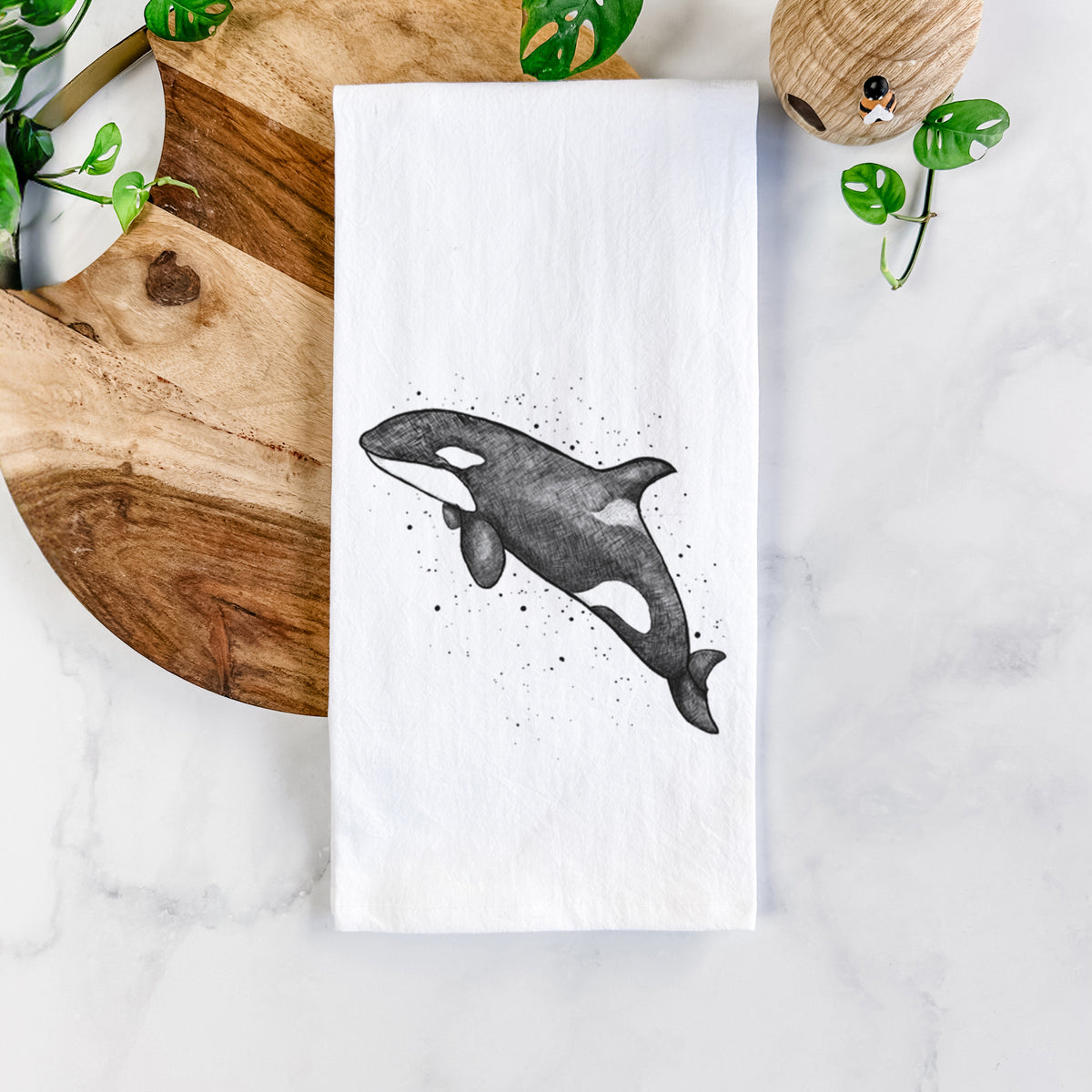 White cotton tea towel printed with a black ink illustration of an orca leaping, with ink splatters, on a marble counter beside a round wooden cutting board and greenery.
