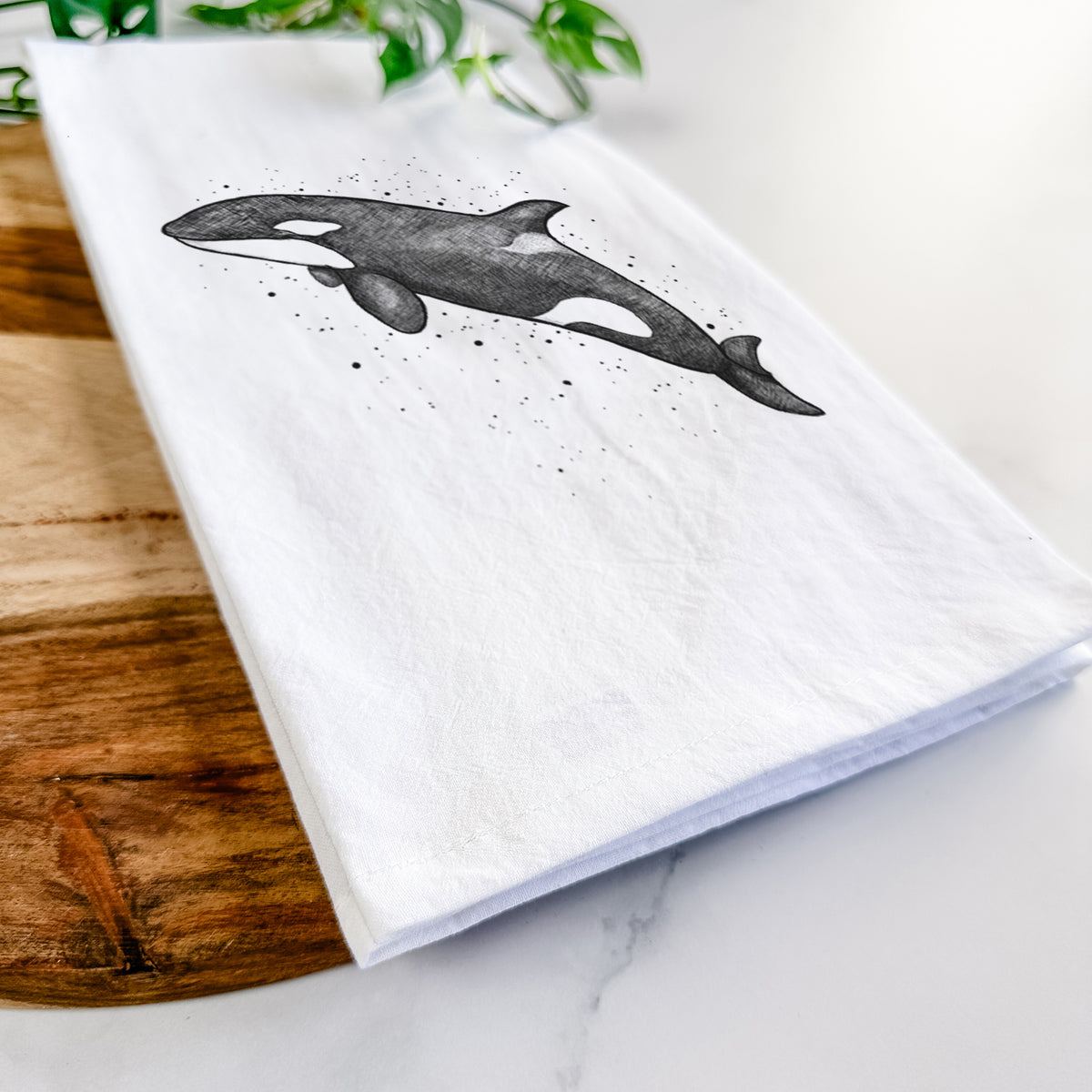 Close-up of a folded white tea towel on a wooden board, showing a black ink illustration of an orca leaping, with ink splatters.