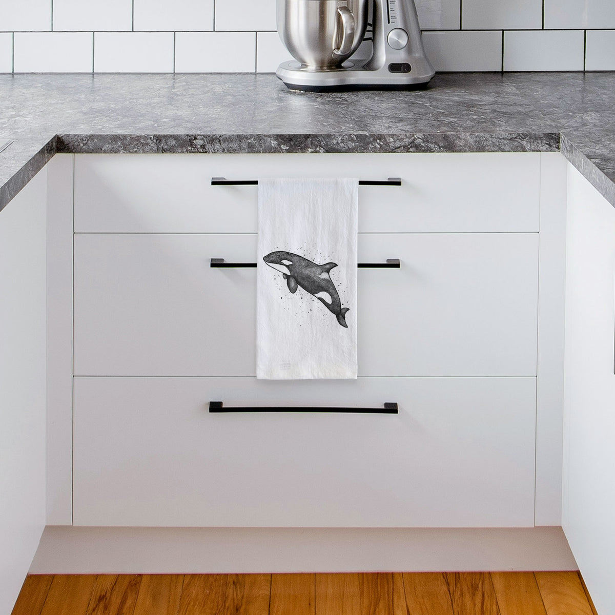 White tea towel printed with a black ink illustration of an orca leaping, with ink splatters, hanging from a drawer handle in a bright white kitchen.