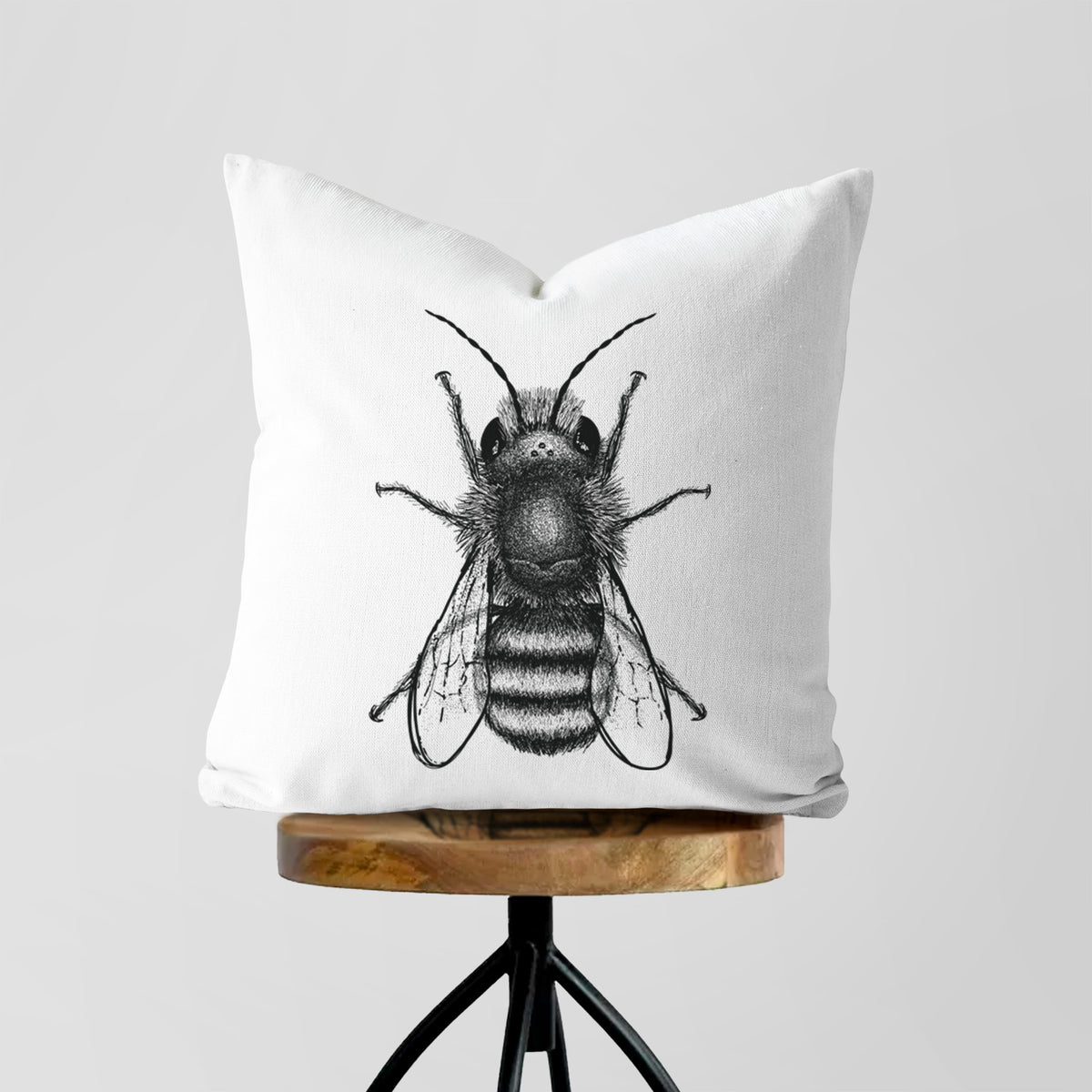 White pillow cover printed with a detailed black ink illustration of a red mason bee with its wings spread, seen from above, on a wooden stool.
