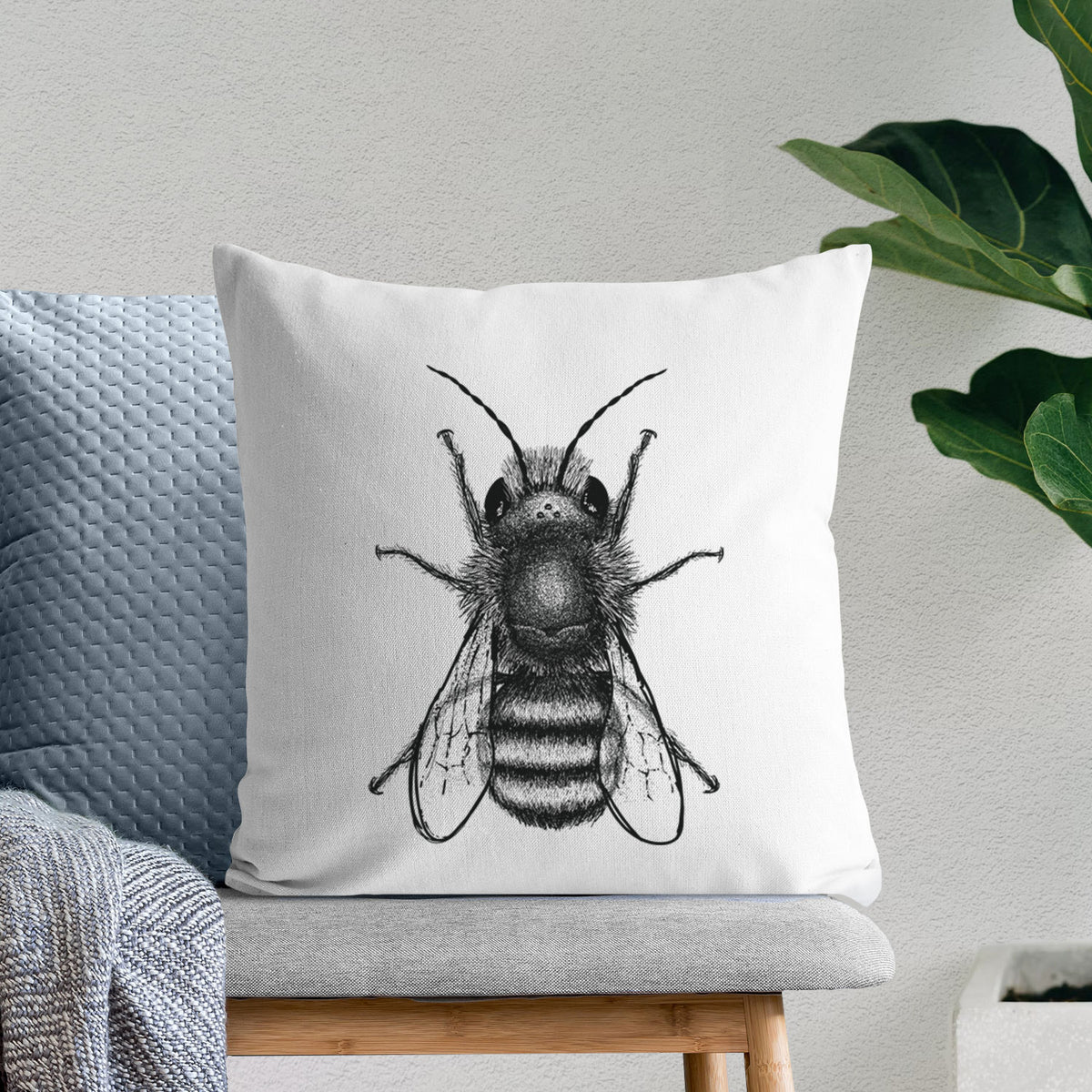 White pillow cover printed with a detailed black ink illustration of a red mason bee with its wings spread, seen from above, on a bench beside plants.