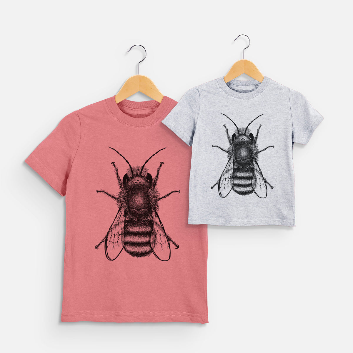Two youth t-shirts in different colors on hangers, printed with a detailed black ink illustration of a red mason bee with its wings spread, seen from above.