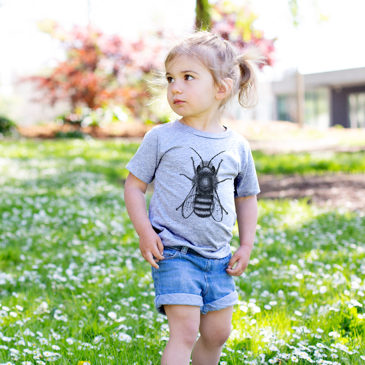 Toddler girl walking outdoors wearing a t-shirt printed with a detailed black ink illustration of a red mason bee with its wings spread, seen from above.