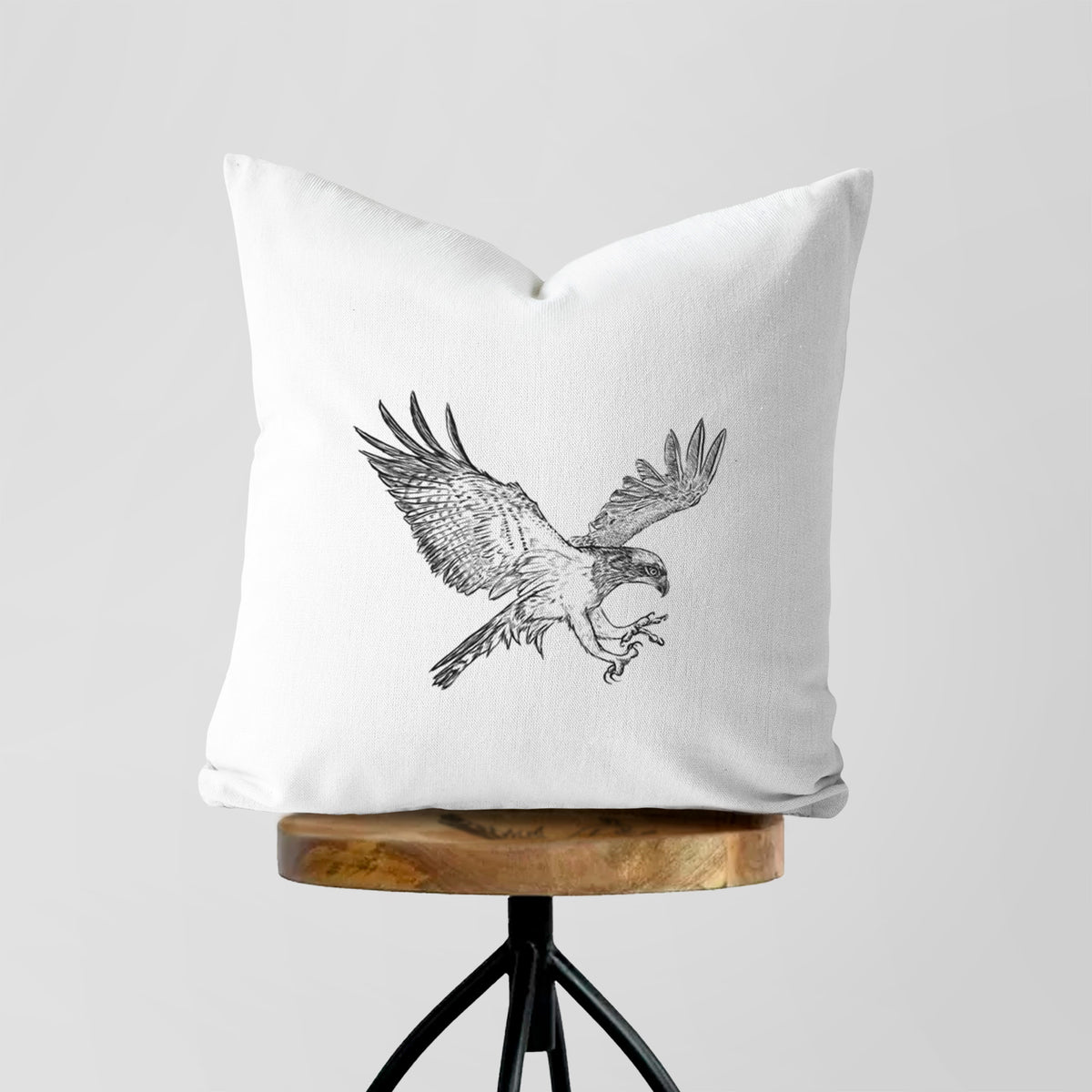 White pillow cover printed with a black ink illustration of an osprey in flight with its wings spread, on a wooden stool.