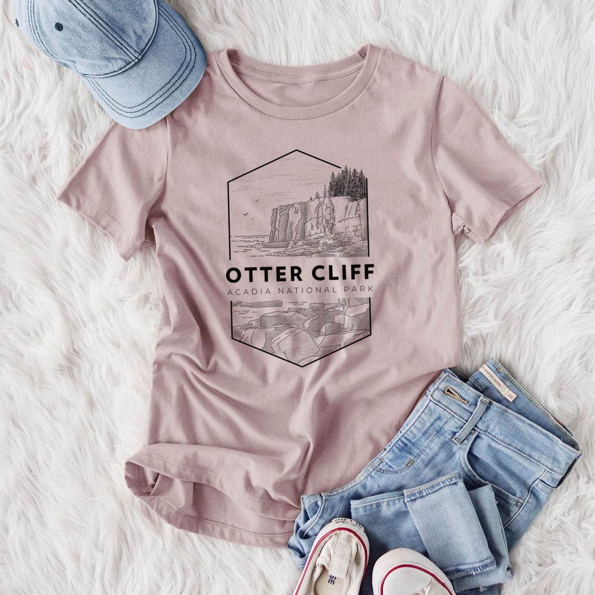 Women's relaxed fit cotton t-shirt printed with a black line art badge of Otter Cliff above the ocean, with the words Otter Cliff, Acadia National Park, styled flat on a white faux fur rug with jeans.