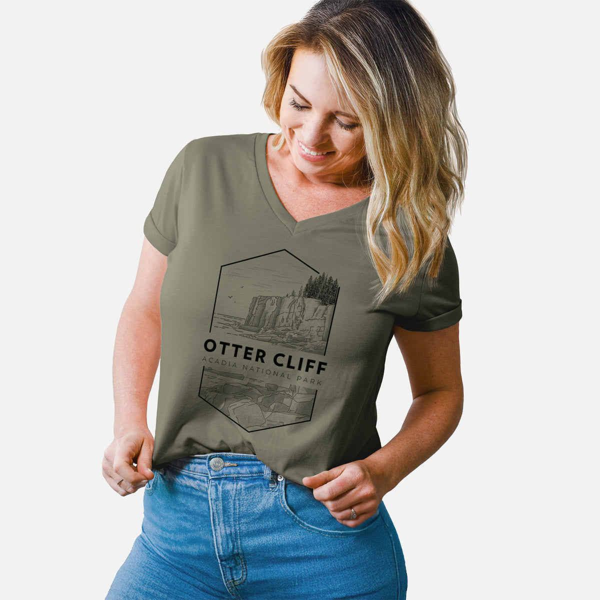 Woman wearing a military green women's relaxed fit cotton V-neck t-shirt printed with a black line art badge of Otter Cliff above the ocean, with the words Otter Cliff, Acadia National Park.