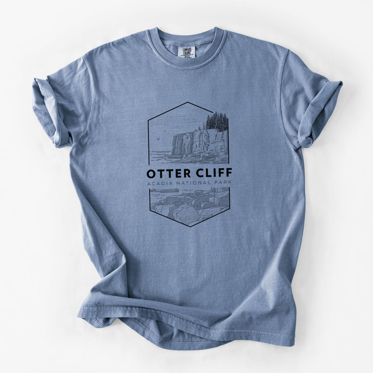 Blue jean Comfort Colors t-shirt laid flat, printed with a black line art badge of Otter Cliff above the ocean, with the words Otter Cliff, Acadia National Park.