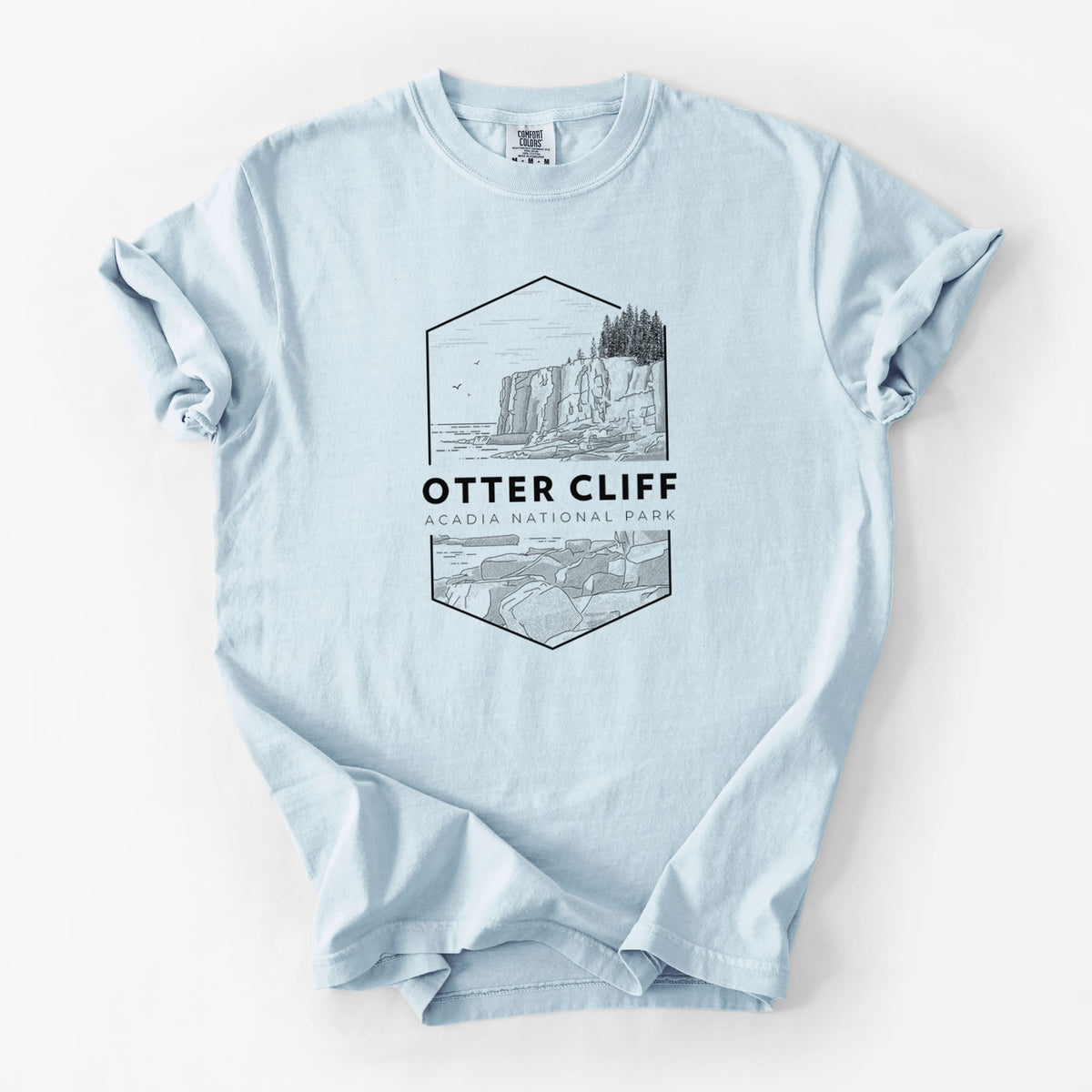 Chambray blue Comfort Colors t-shirt laid flat, printed with a black line art badge of Otter Cliff above the ocean, with the words Otter Cliff, Acadia National Park.