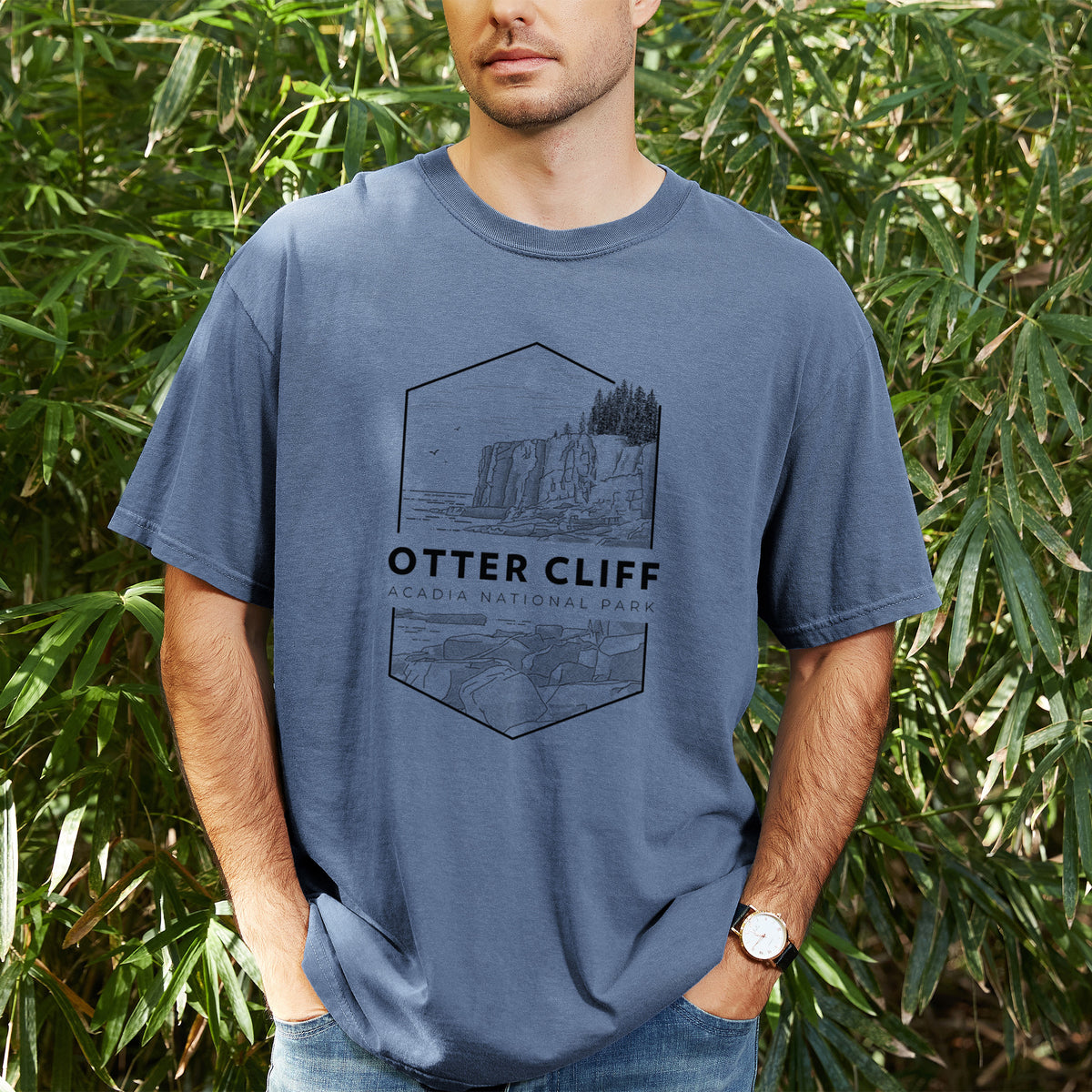 Man wearing a Comfort Colors t-shirt printed with a black line art badge of Otter Cliff above the ocean, with the words Otter Cliff, Acadia National Park, outdoors.
