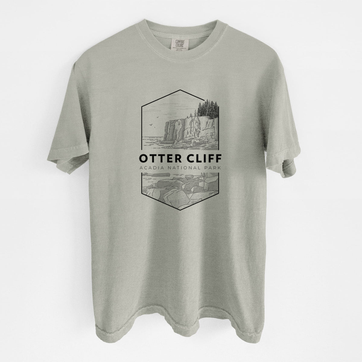 Sandstone beige Comfort Colors t-shirt laid flat, printed with a black line art badge of Otter Cliff above the ocean, with the words Otter Cliff, Acadia National Park.