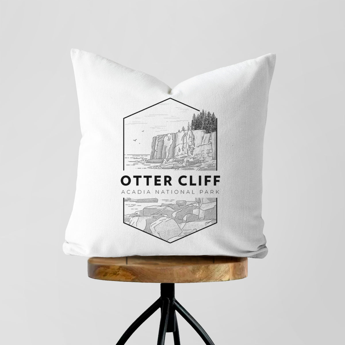 White pillow cover printed with a black line art badge of Otter Cliff above the ocean, with the words Otter Cliff, Acadia National Park, on a wooden stool.