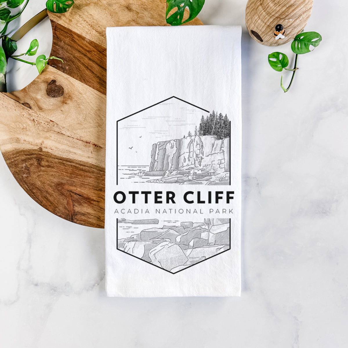 White cotton tea towel printed with a black line art badge of Otter Cliff above the ocean, with the words Otter Cliff, Acadia National Park, on a marble counter beside a round wooden cutting board and greenery.