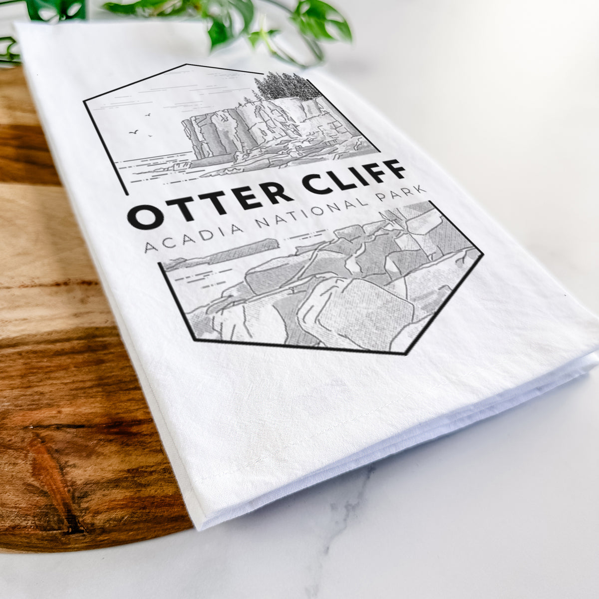 Close-up of a folded white tea towel on a wooden board, showing a black line art badge of Otter Cliff above the ocean, with the words Otter Cliff, Acadia National Park.