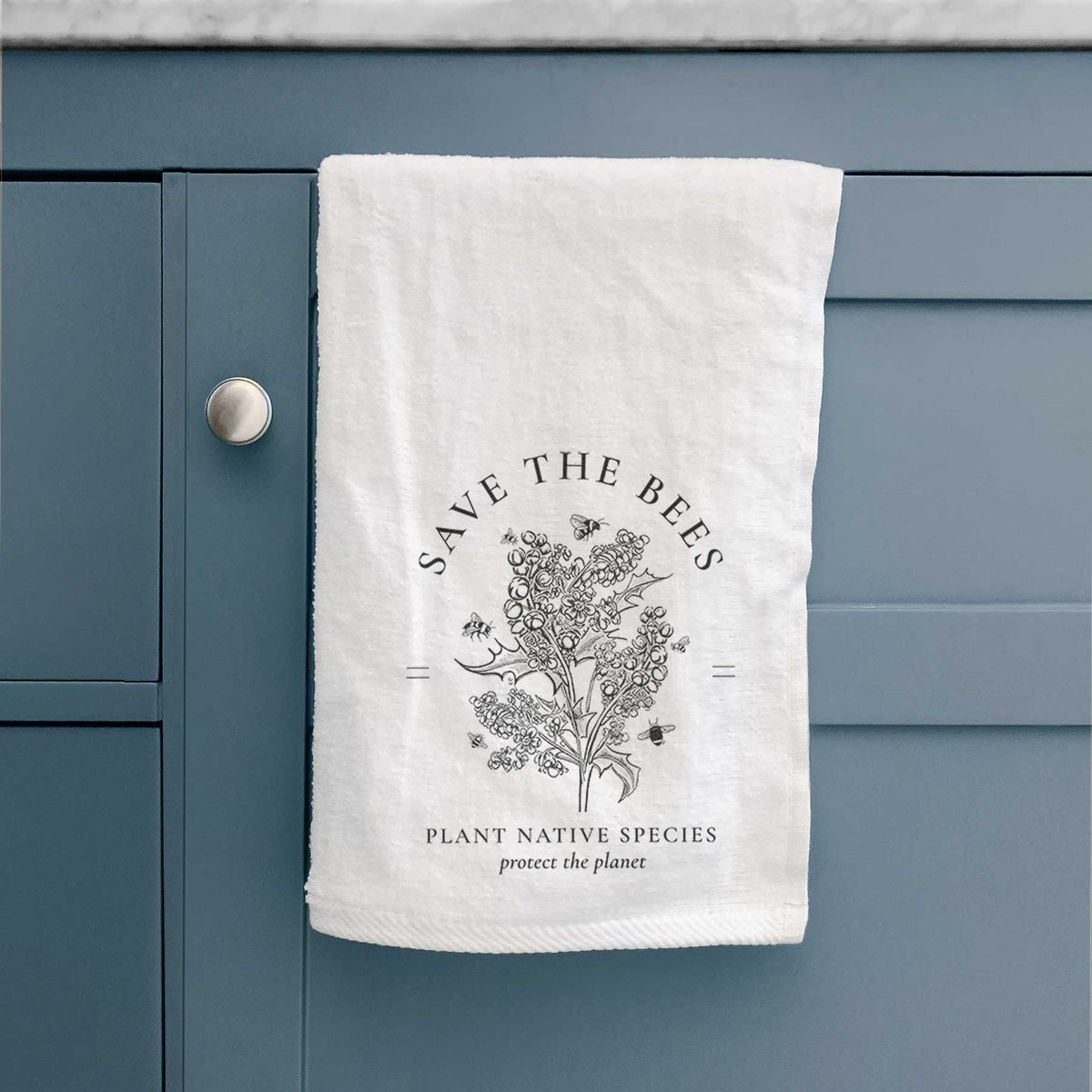 White hand towel printed with a black ink wildflower bouquet with bees, framed by the words Save the Bees, Plant Native Species, hanging on a cabinet door.