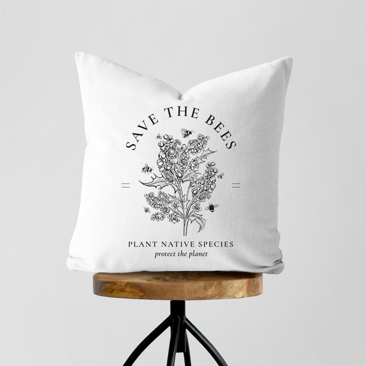 White pillow cover printed with a black ink wildflower bouquet with bees, framed by the words Save the Bees, Plant Native Species, on a wooden stool.