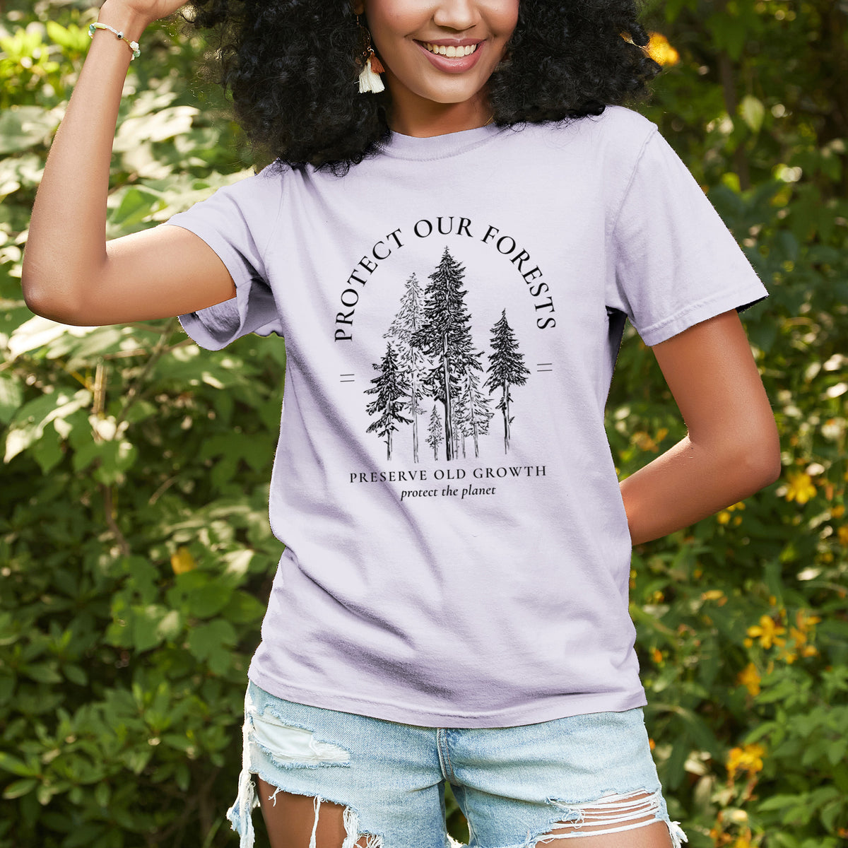 Woman wearing a Comfort Colors t-shirt printed with a black ink illustration of a stand of tall pine trees, framed by the words Protect Our Forests, Preserve Old Growth, outdoors.