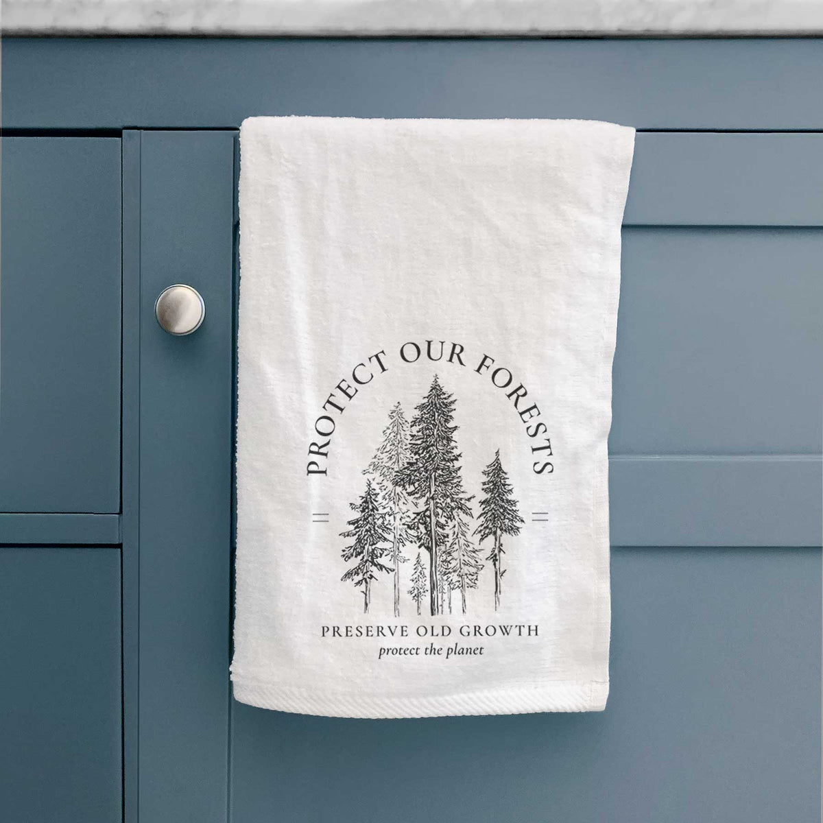 White hand towel printed with a black ink illustration of a stand of tall pine trees, framed by the words Protect Our Forests, Preserve Old Growth, hanging on a cabinet door.