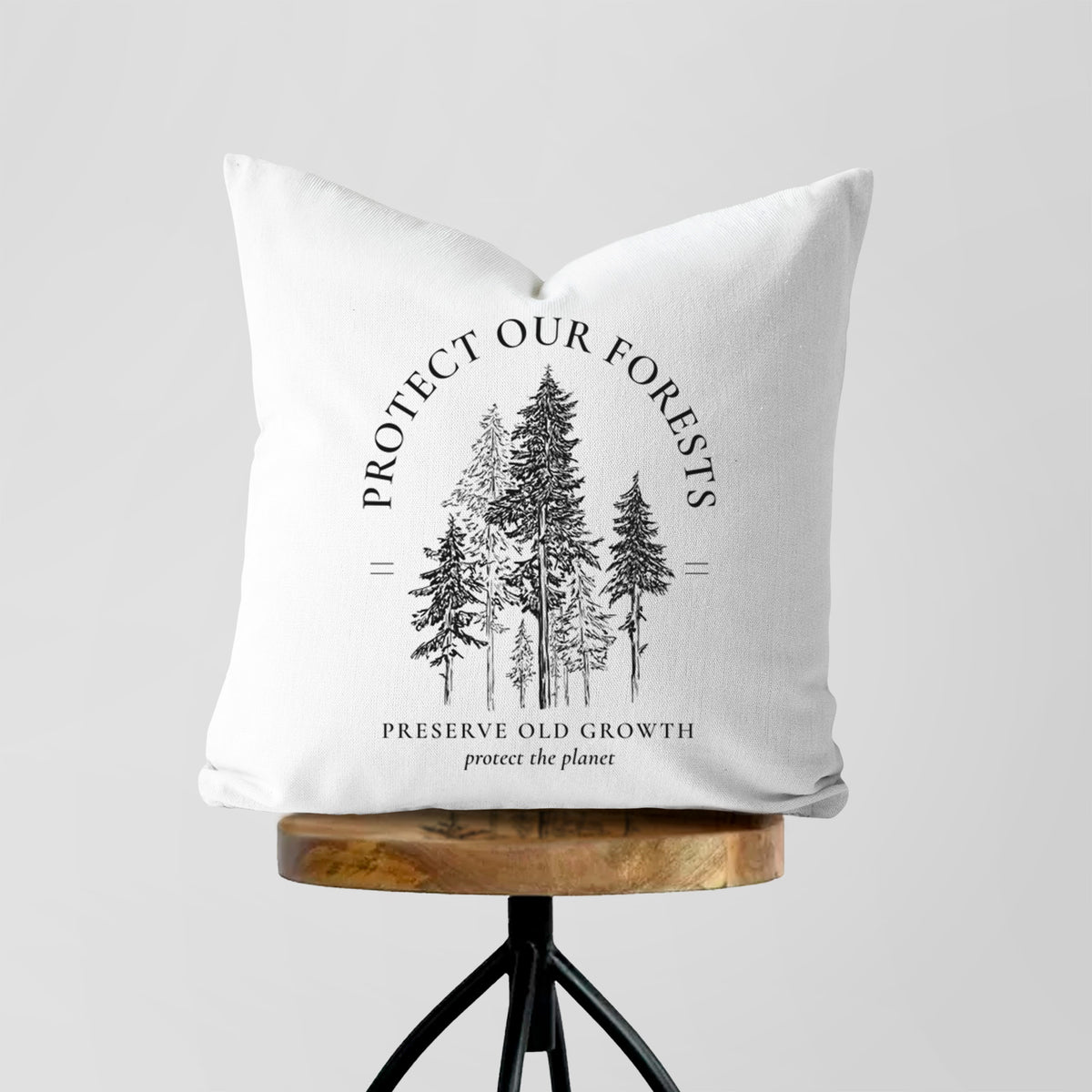 White pillow cover printed with a black ink illustration of a stand of tall pine trees, framed by the words Protect Our Forests, Preserve Old Growth, on a wooden stool.